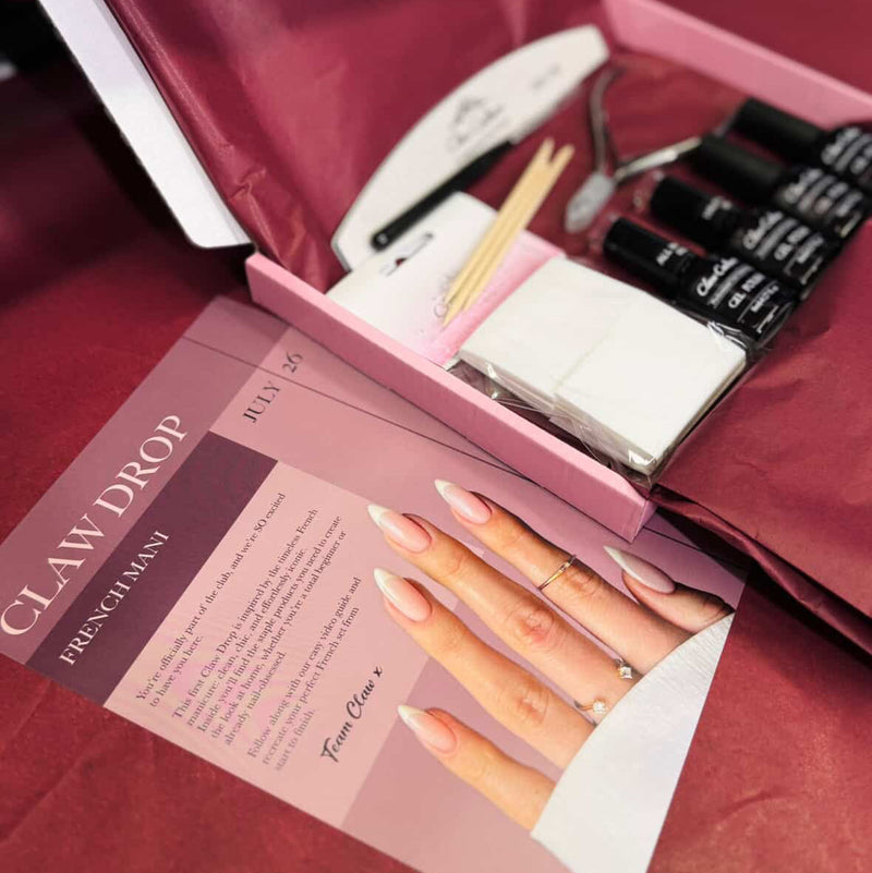 A nail kit displayed in an open pink box on a burgundy surface, including black nail polish bottles, white nail files, wooden sticks, and a printed instruction card titled 'CLAW DROP FRENCH MANI'.