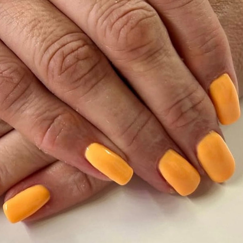 Close-up of hands with orange nail polish on a neutral background, featuring a brand logo in the corner.