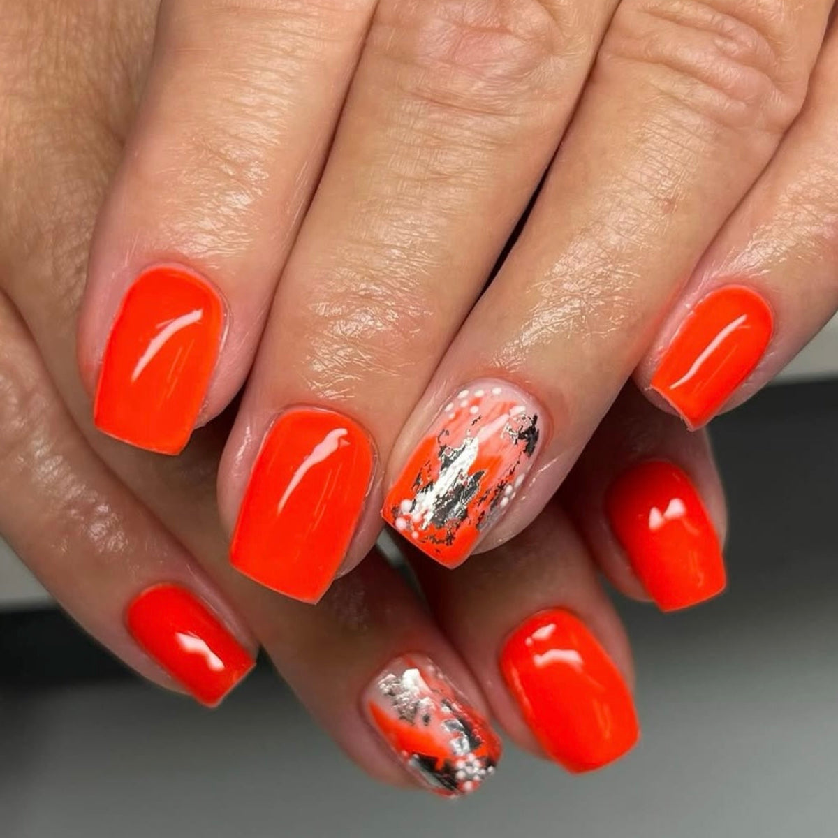 Close-up of a hand with bright orange nail polish and a silver leaf design on a neutral background